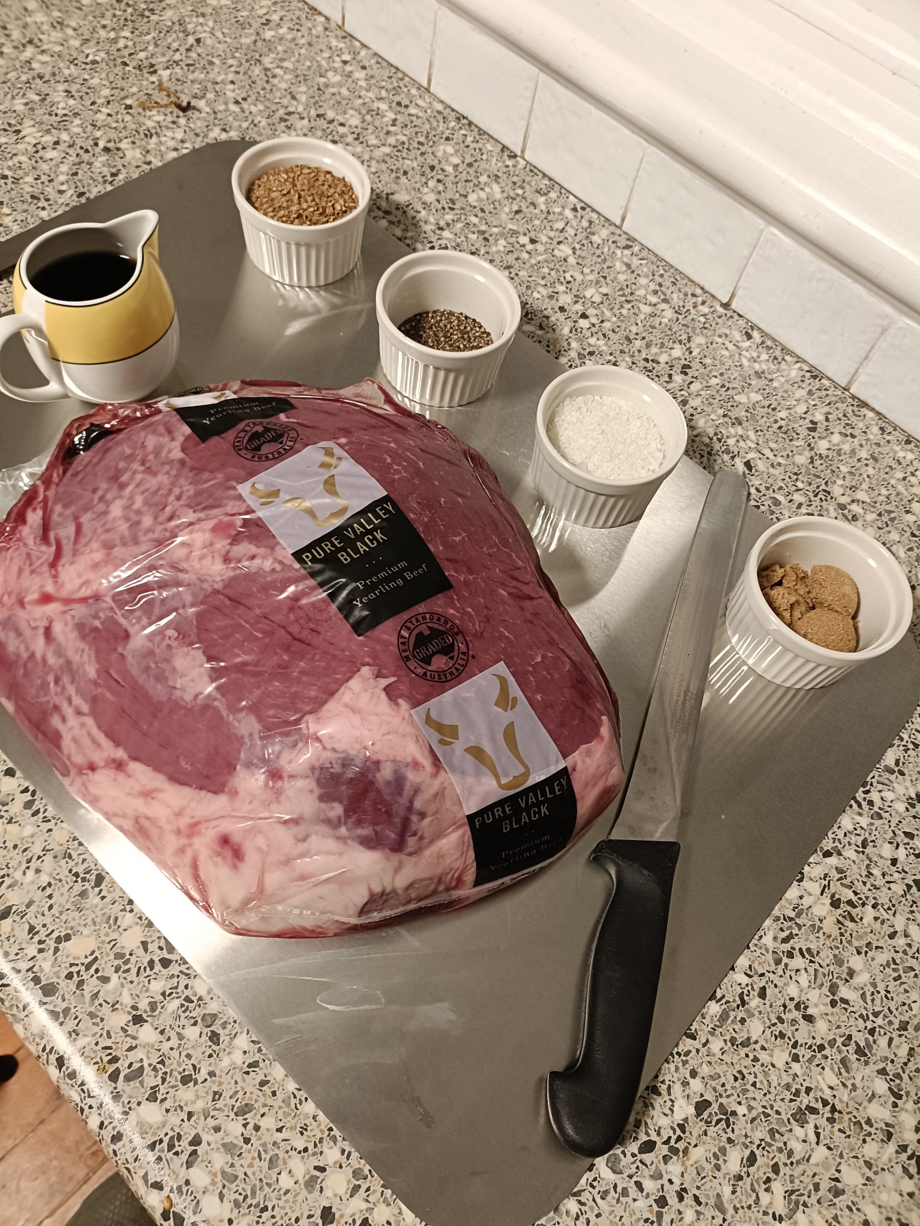 A whole beef cut beside ramekins of whole coriander, cracked pepper, salt and sugar, ready for curing.